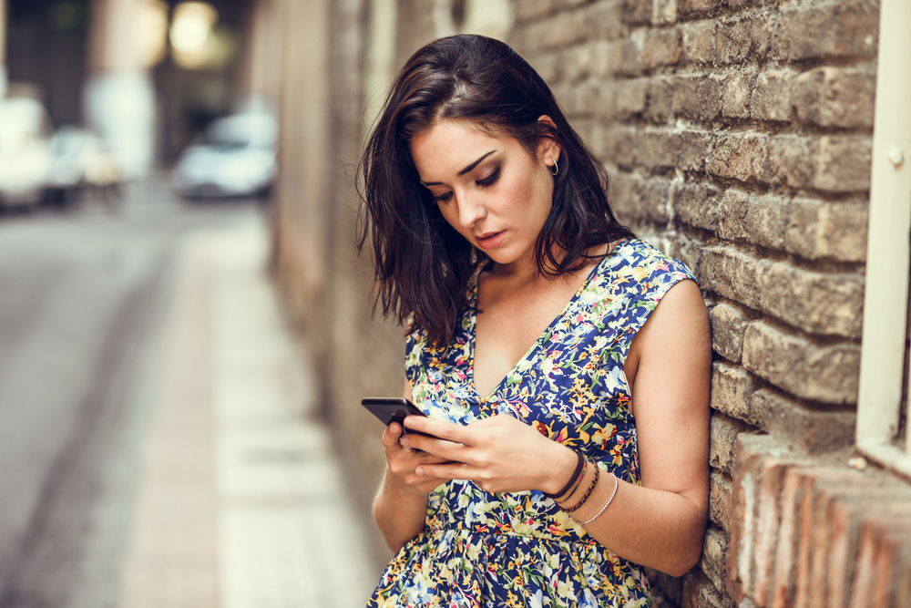 Serious young woman using her smart phone outdoors. Girl wearing flower dress in urban background. Technology concept.