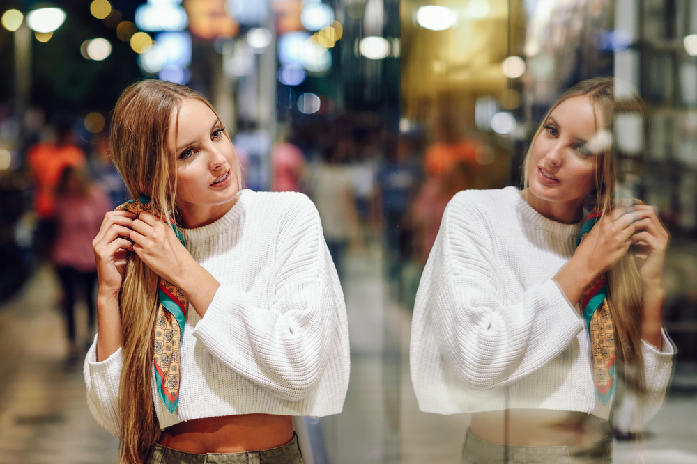 Blonde girl wearing white sweater standing in the street with defocused city lights at the background. Pretty woman with pigtail hairstyle at night reflected in a shop window.