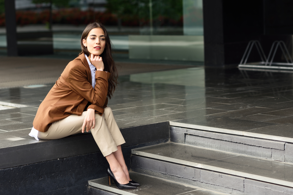 Young businesswoman sitting outside of office building. Beautiful woman wearing formal wear. Young girl with brown jacket and trousers in urban background.