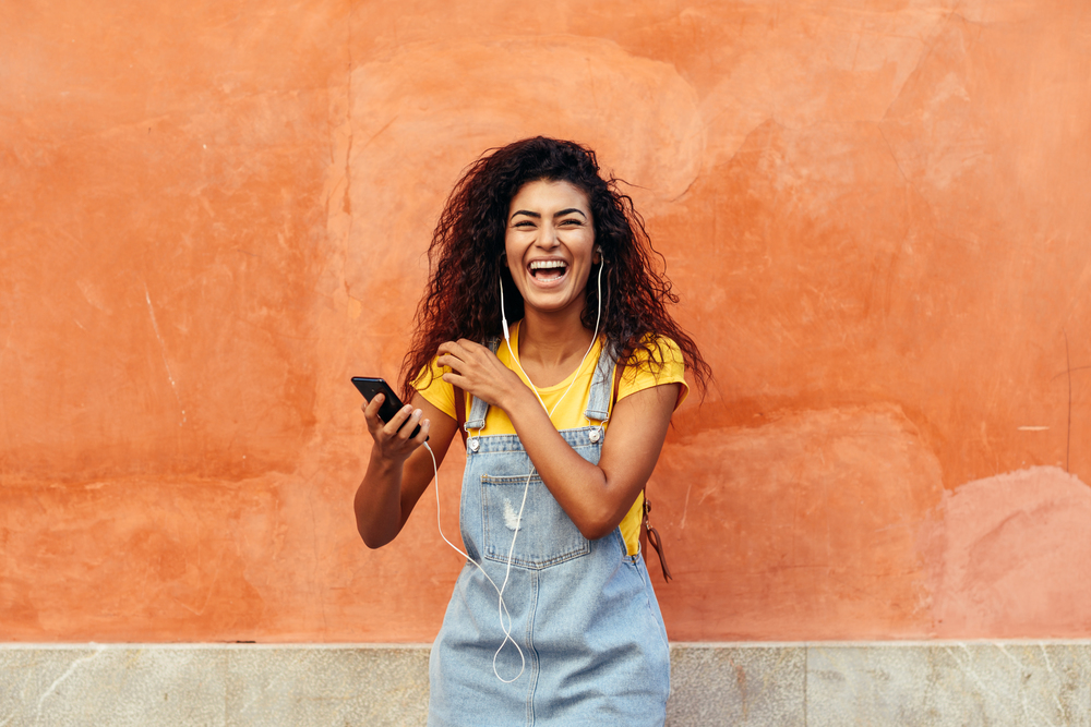 Young black woman laughing and listening to music with earphones outdoors. Arab girl in casual clothes with curly hairstyle in urban background.