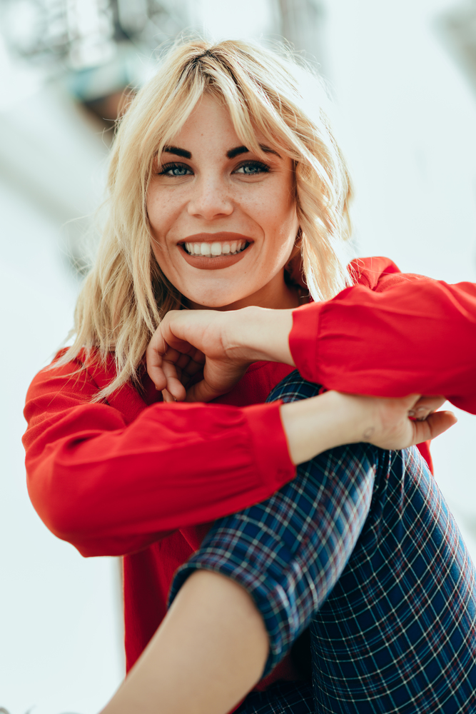 Happy young blond woman sitting on urban background. Smiling blonde girl with red shirt enjoying life outdoors.