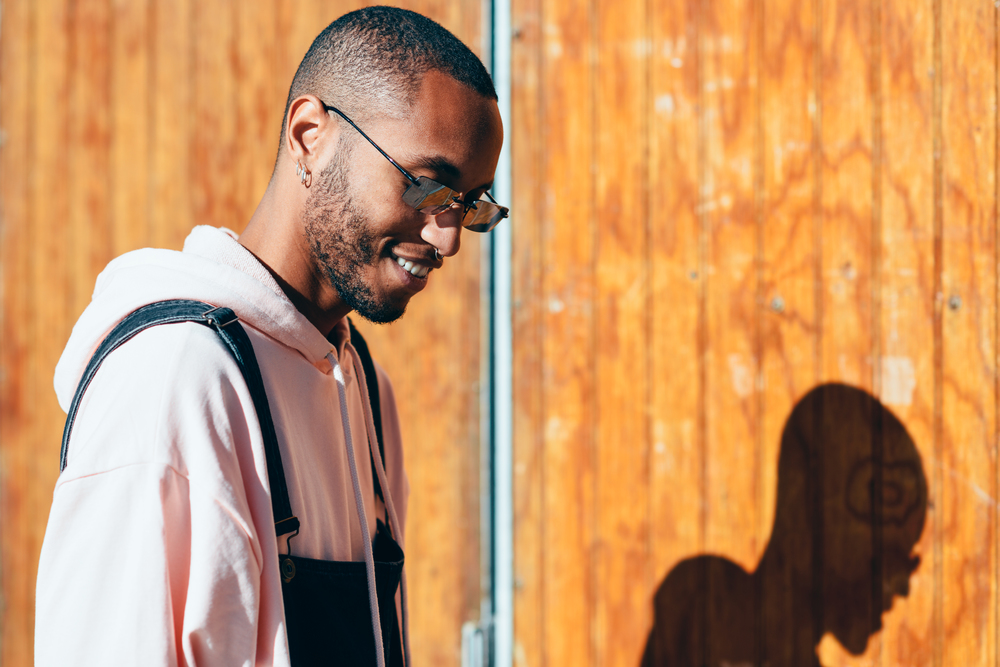 Young black man wearing casual clothes and sunglasses, smiling against a wooden background. Millennial african guy with bib pants outdoors