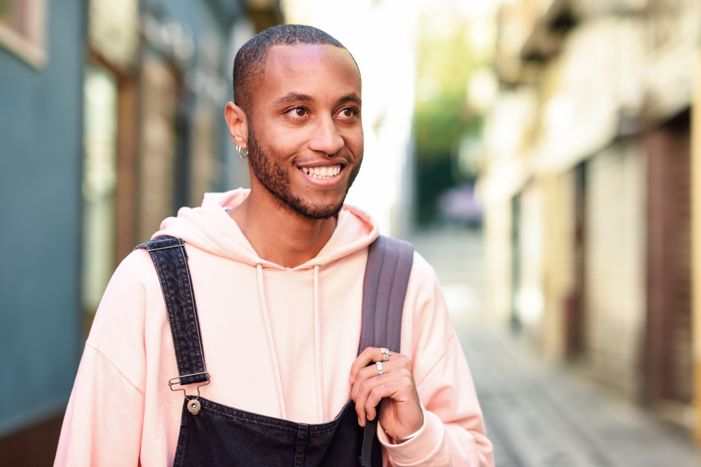 Young black man wearing casual clothes walking smiling down the street. Millennial african guy with bib pants outdoors