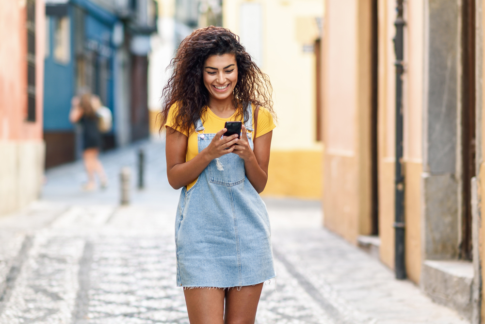 Young North African woman walking on the street looking at her smart phone. Smiling Arab girl in casual clothes with black curly hairstyle.