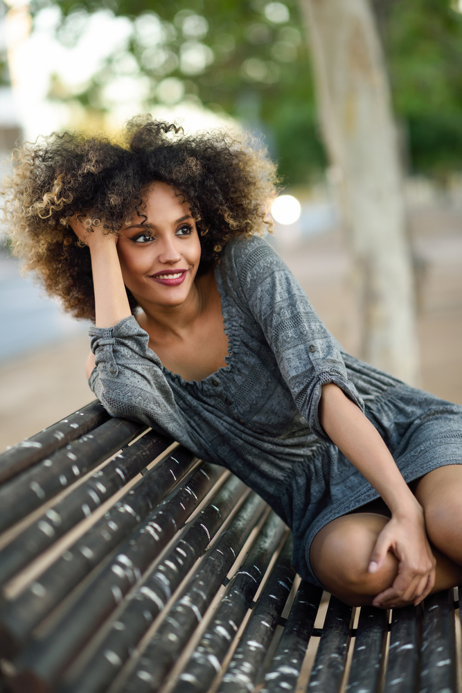 Funny black woman with afro hairstyle sitting on a bench in urban background. Mixed girl wearing casual clothes. Young black woman with afro hairstyle smiling in urban background