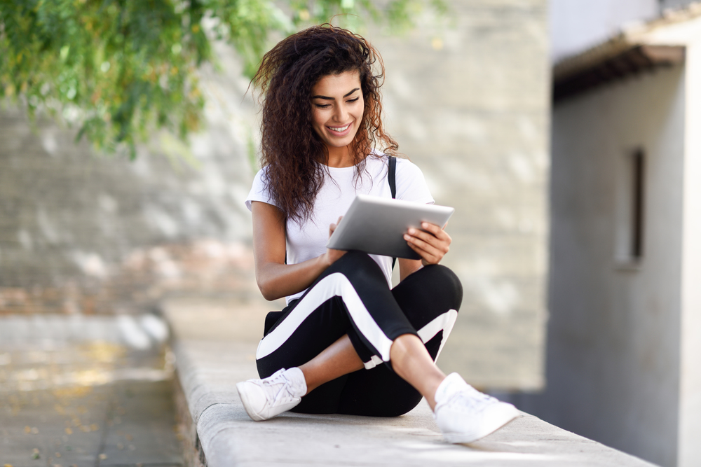 Young North African woman with looking at her digital tablet outdoors. Arab girl wearing sportswear urban background.. Young black woman in sportswear using digital tablet outdoors