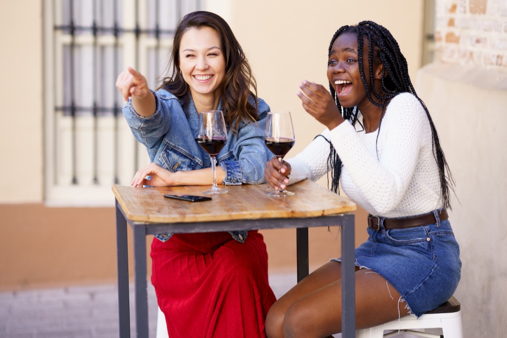 Two friends drinking red wine sitting at a table outside a bar. Multiethnic women.. Two women drinking red wine sitting at a table outside a bar.