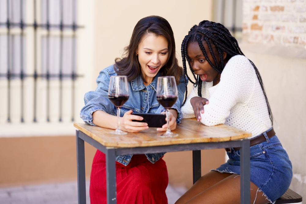 Two women looking at their smartphone together while having a glass of wine on the terrace of a bar. Multiethnic women.. Two women looking at their smartphone together while having a glass of wine.