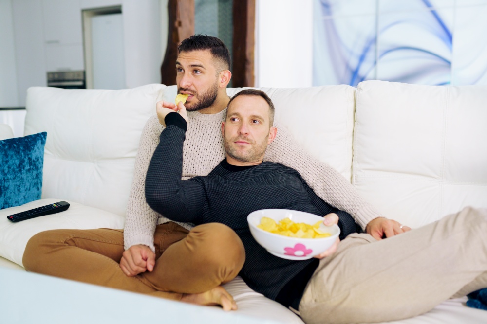 Gay couple sitting on the couch at home watching something on TV and having a snack. Homosexual relationship concept.. Gay couple sitting on the couch at home watching something on TV and having a snack.