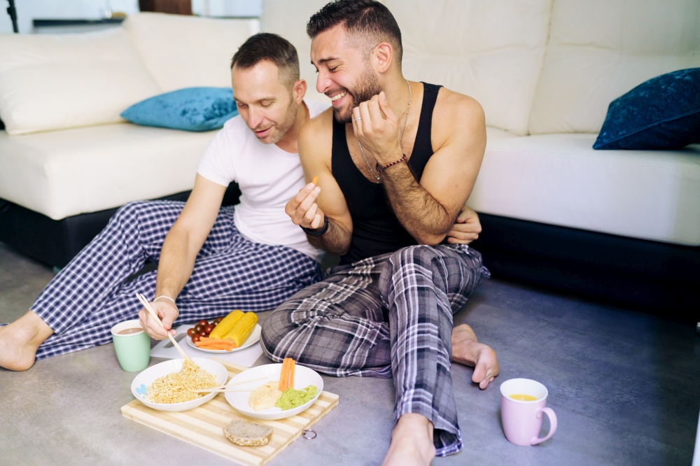 Gay couple eating together sitting on their living room floor. Homosexual lifestyle concept.. Gay couple eating together sitting on their living room floor.