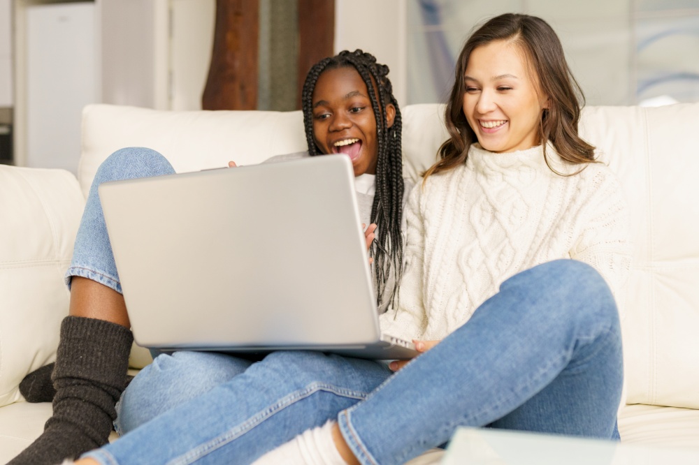 Two female student friends sitting on the couch at home using a laptop. Multiethnic women.. Two female student friends sitting on the couch at home using a laptop.