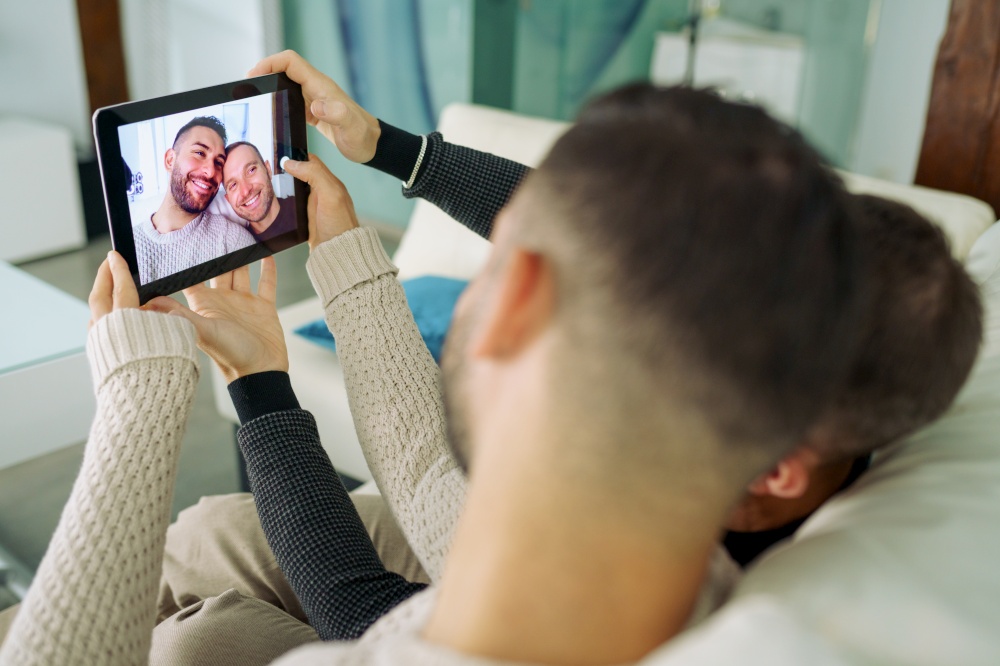 Gay couple making a selfie with a digital tablet sitting on the couch at home. Homosexual Lifestyle concept.. Gay couple making a selfie with a digital tablet sitting on the couch at home.