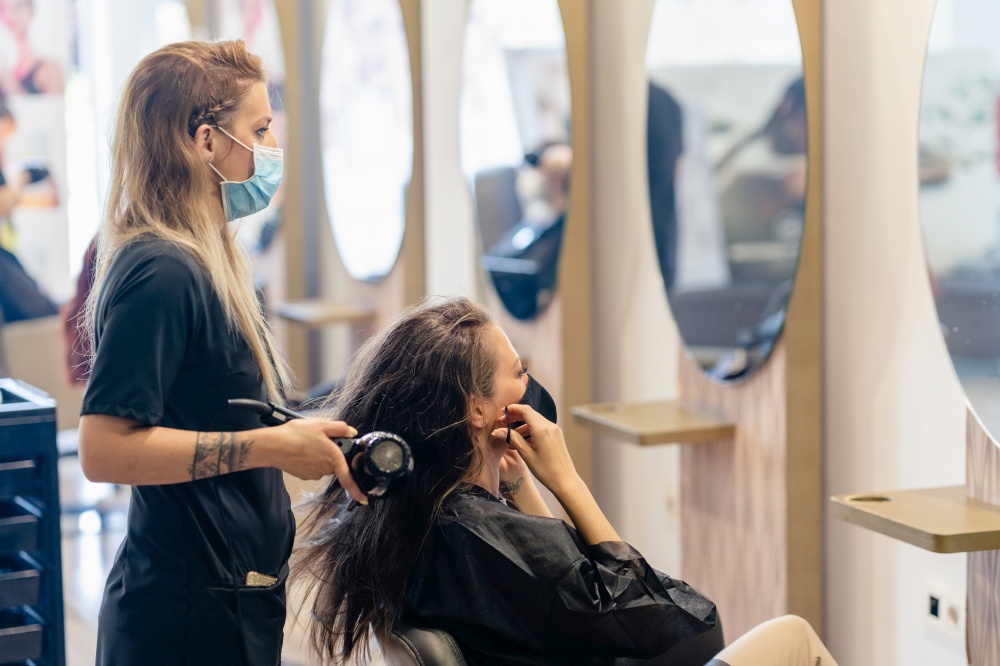 Female hairdresser drying her client&rsquo;s hair with a hairdryer, wearing protective masks in a beauty centre. Business and beauty concepts. Hairdresser drying her client&rsquo;s hair with a hairdryer wearing protective masks in a beauty centre.