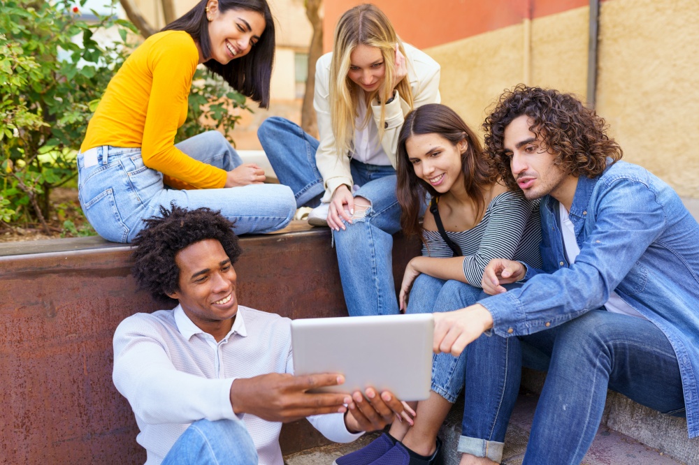 Multi-ethnic group of young people looking at a digital tablet outdoors in urban background. Group of men and woman sitting together on steps.. Multi-ethnic group of young people looking at a digital tablet outdoors in urban background.