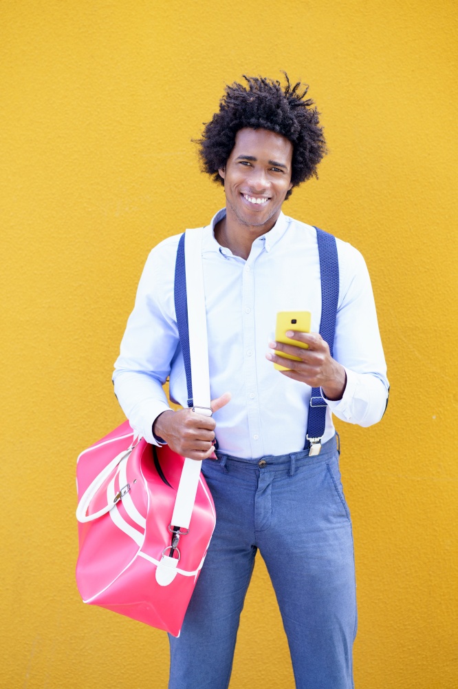 Black man with afro hairstyle carrying a sports bag and smartphone against a yellow urban background. Guy with curly hair wearing shirt and suspenders.. Black man with afro hairstyle carrying a sports bag and smartphone in yellow background.