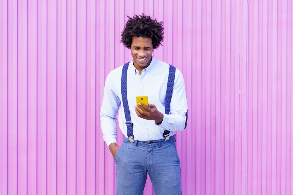 Black man with afro hairstyle using a smartphone against a pink blinds background. Guy with curly hair wearing shirt and suspenders.. Black man with afro hairstyle carrying a sports bag and smartphone in yellow background.