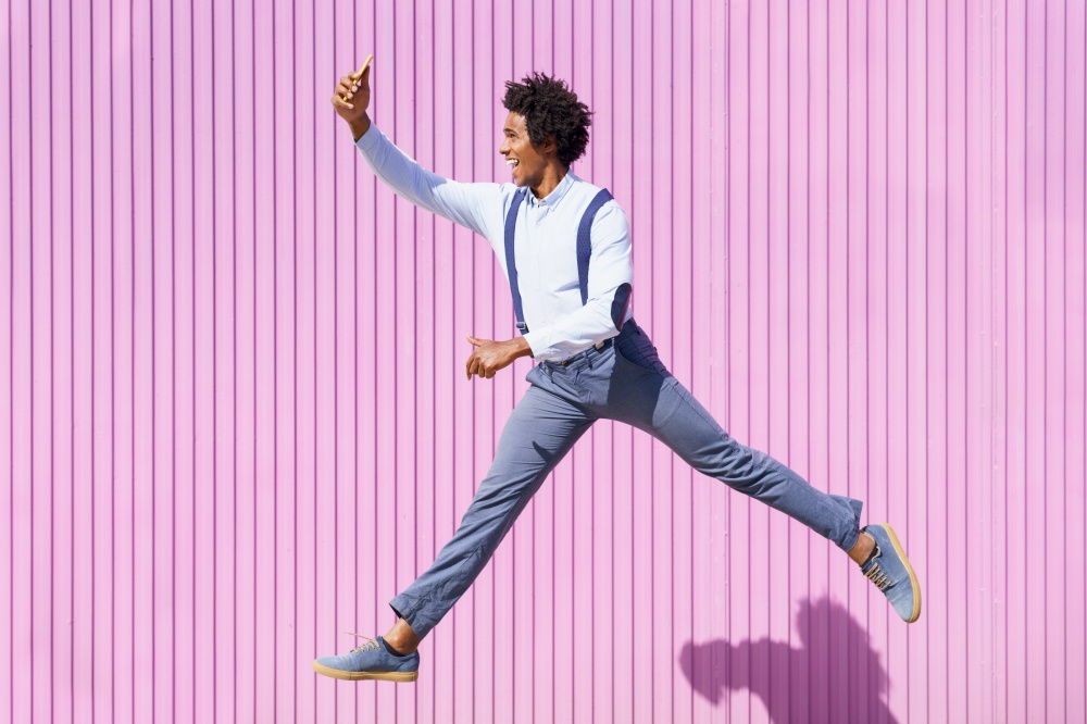 Black man, with afro hair, taking a selfie with his smartphone while jumping on a background of pink shutters.. Black man taking a selfie with his smartphone while jumping outdoors.
