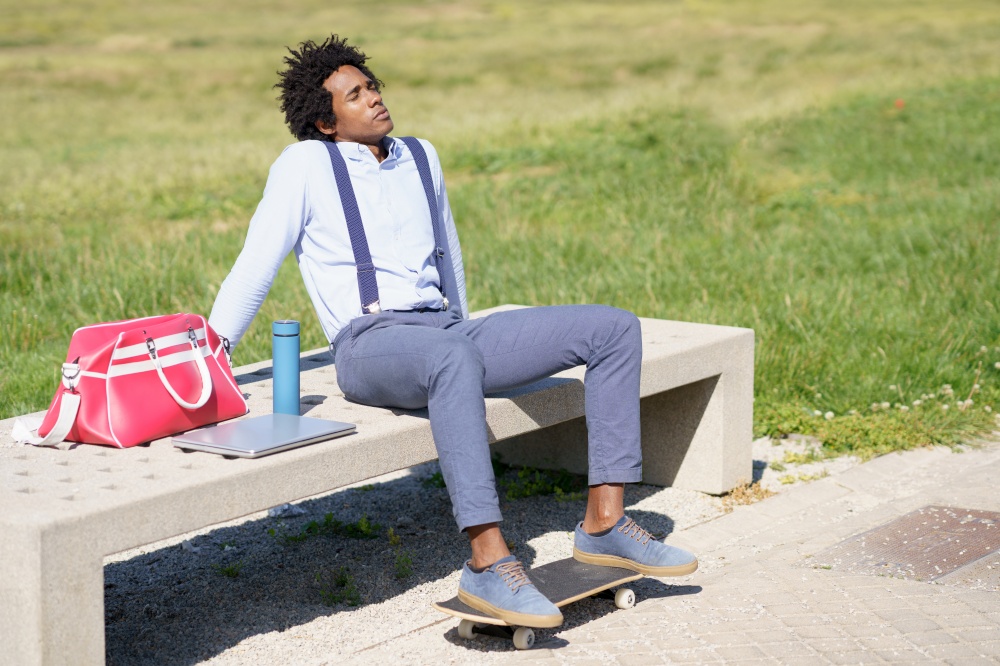 Black businessman with afro hair taking a coffee break sitting on a park bench with his feet on his skateboard.. Black man with afro hair taking a coffee break sitting on a park bench.