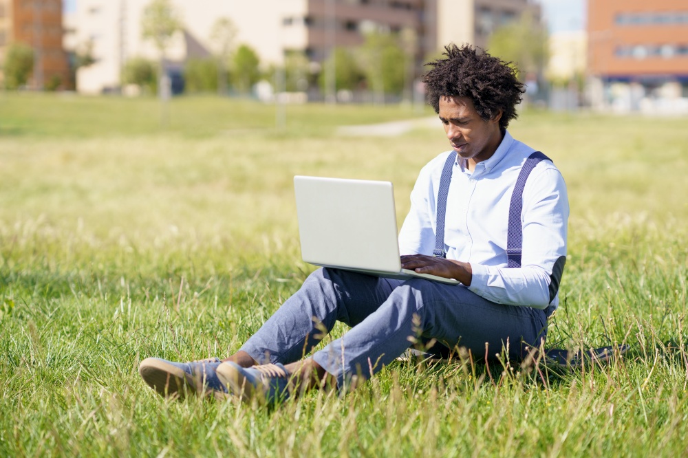Black working man with afro hair using his laptop sitting on skateboard on the grass of an urban park.. Black man with afro hair using his laptop sitting on skateboard on the grass of an urban park.