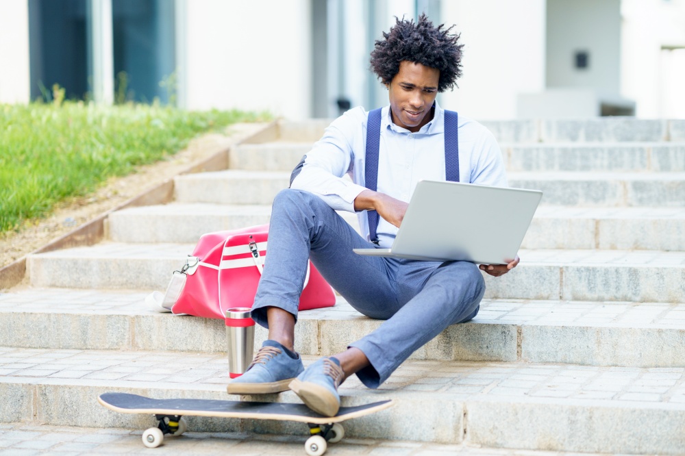 Black businessman with afro hair and skateboard using his laptop computer sitting on some steps of an office building.. Black businessman with afro hair and skateboard using his laptop computer sitting on some steps.