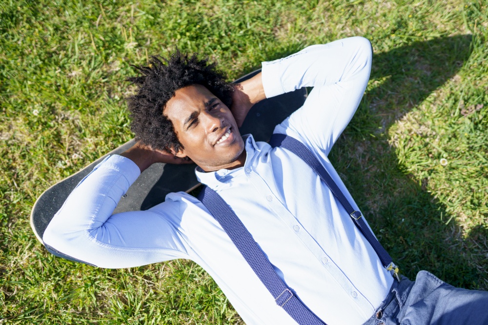 Black man worker with afro hair resting lying on the grass with his skateboard. Black businessman resting lying on the grass with his skateboard