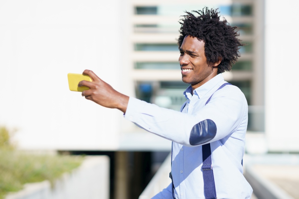 Black businessman with afro hairstyle taking a selfie with his smartphone near an office building.. Black businessman taking a selfie with his smartphone near an office building.