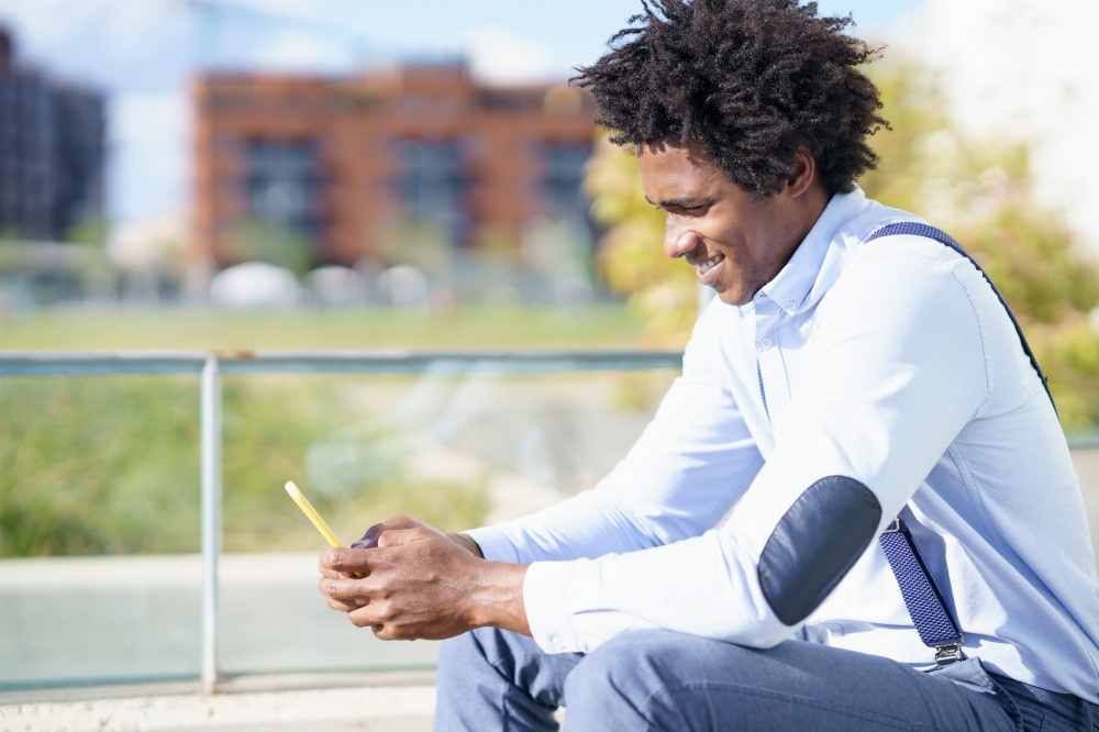 Black man with afro hairstyle using a smartphone near an office building. Guy with curly hair wearing shirt and suspenders.. Black man with afro hairstyle using a smartphone sitting near an office building.