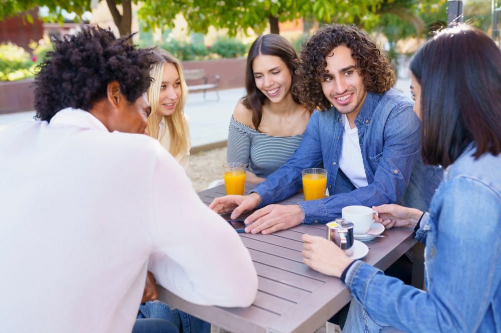Young man showing something to his multi-ethnic group of friends on his smartphone, while having drinks at an outdoor table in a bar.. Guy showing his smartphone to his group of friends while having drinks at an outdoor bar