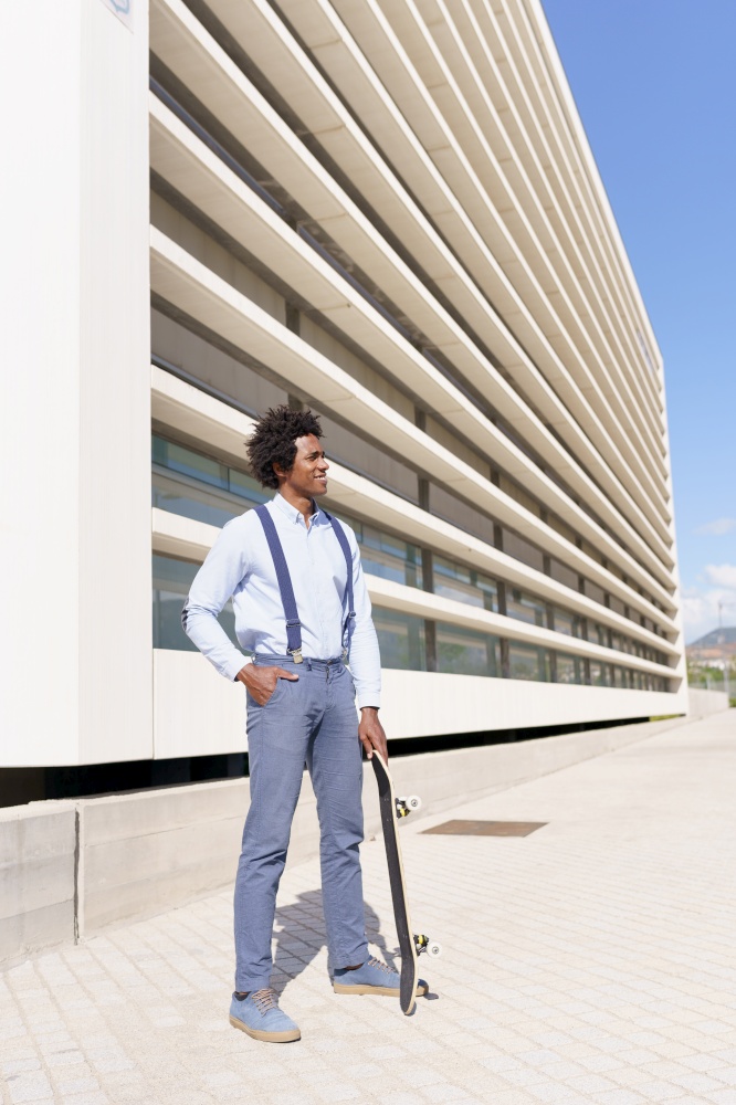 Black businessman worker with afro hairstyle standing next to an office building with a skateboard.. Black male worker standing next to an office building with a skateboard.
