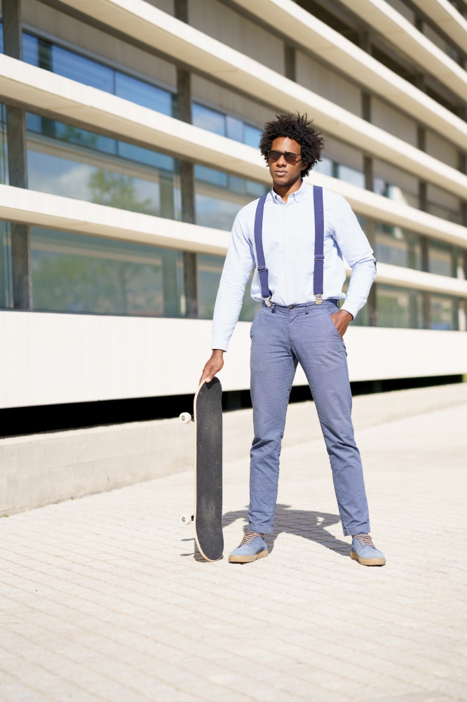 Black male worker with afro hairstyle standing next to an office building with a skateboard and sunglasses.. Black male worker standing next to an office building with a skateboard.