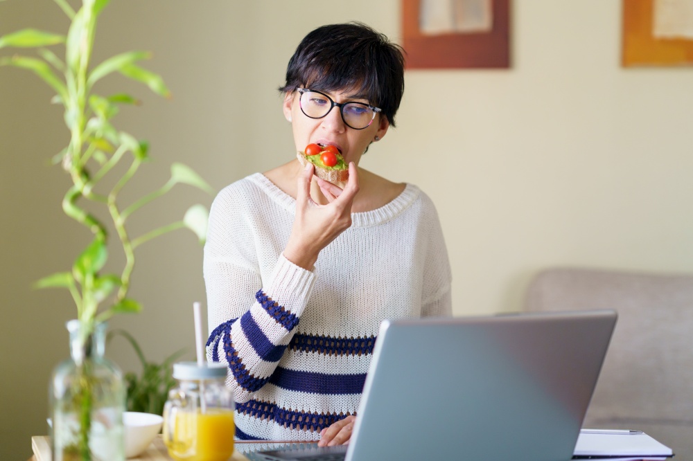 Woman eating some healthy food, while teleworking from home on her laptop. Female in her 50s. Woman eating some healthy food, while teleworking from home on her laptop.