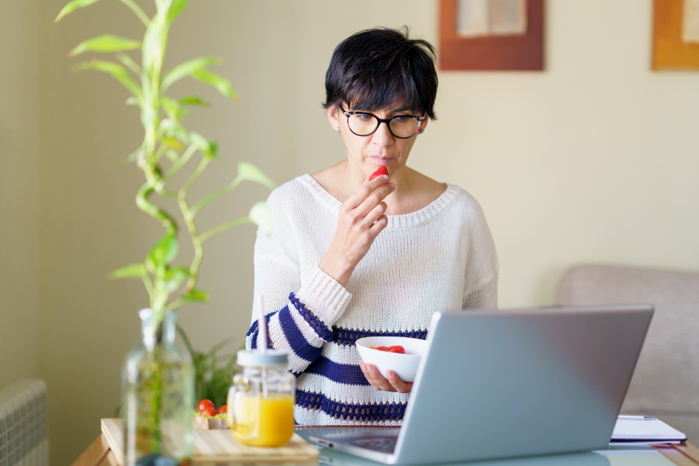 Woman eating strawberries while teleworking from home on her laptop. Female in her 50s. Woman eating strawberries while teleworking from home on her laptop