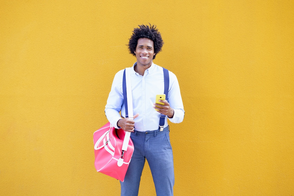 Guy with curly hair wearing shirt and suspenders. Black man with afro hairstyle carrying a sports bag and smartphone against a yellow urban background.. Black man with afro hairstyle carrying a sports bag and smartphone in yellow background.