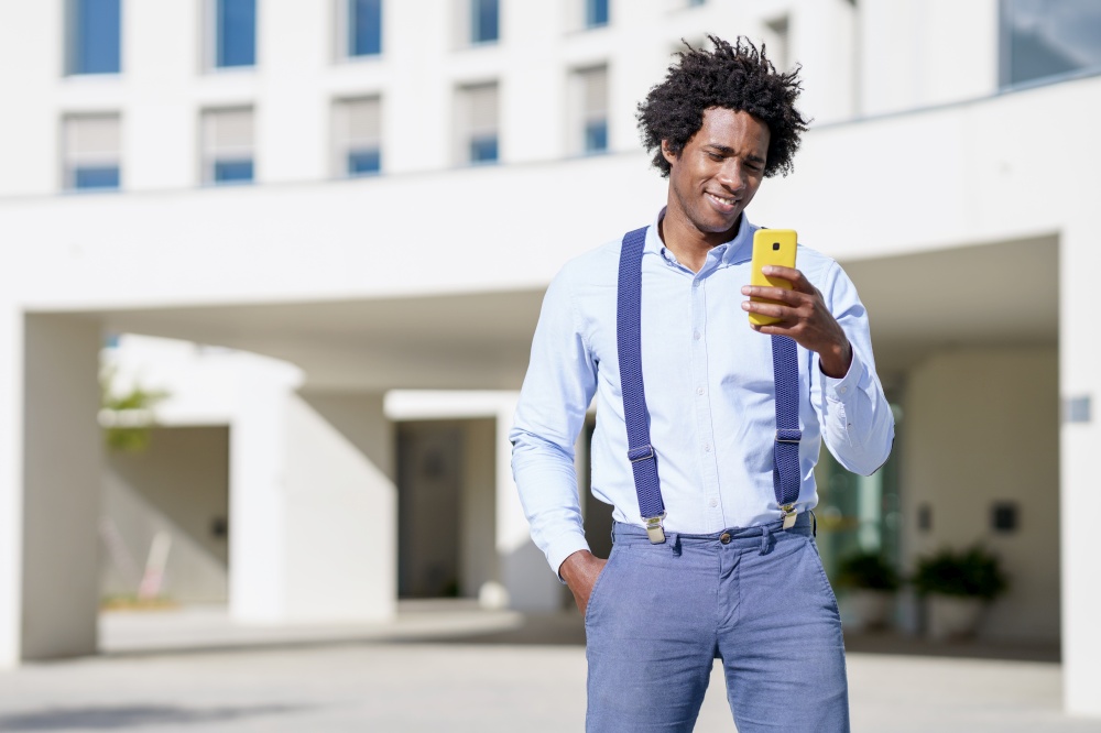 Black man with afro hairstyle using a smartphone near an office building. Guy with curly hair wearing shirt and suspenders.. Black man with afro hairstyle using a smartphone near an office building.
