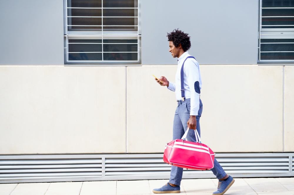 Black man with afro hairstyle carrying a sports bag and smartphone in urban background. Guy with curly hair wearing shirt and suspenders.. Black man with afro hairstyle carrying a sports bag and smartphone outdoors.