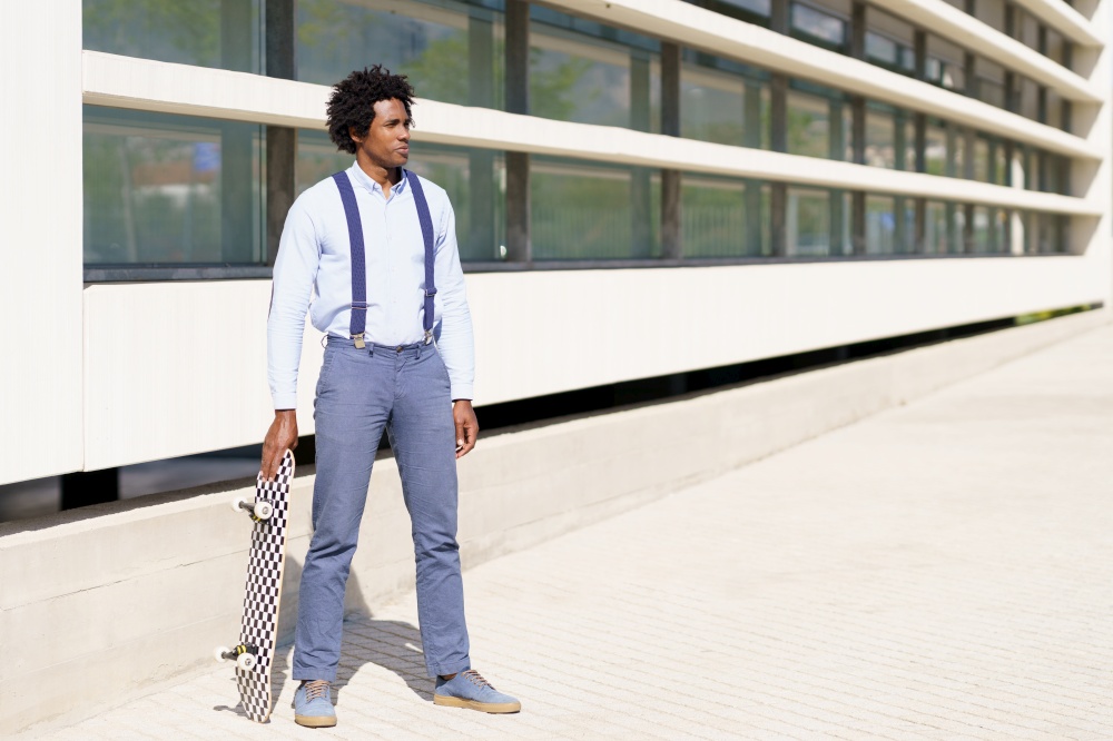 Black businessman worker with afro hairstyle standing next to an office building with a skateboard.. Black male worker standing next to an office building with a skateboard.