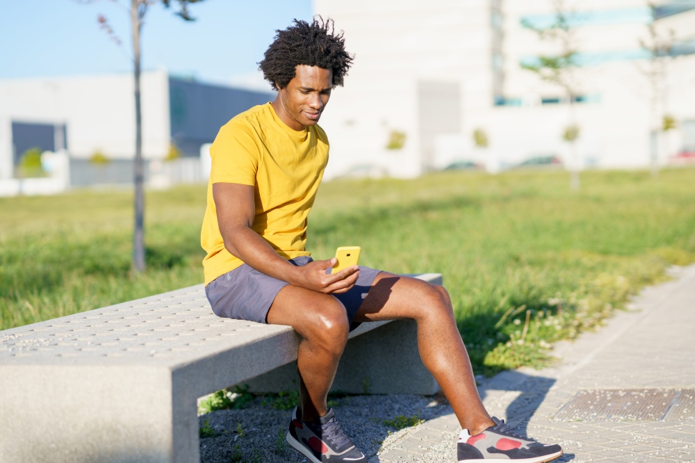 Black man with afro hair and earphones consulting his smartphone with some exercise app while resting from his workout.. Black man consulting his smartphone with some exercise app while resting from his workout.
