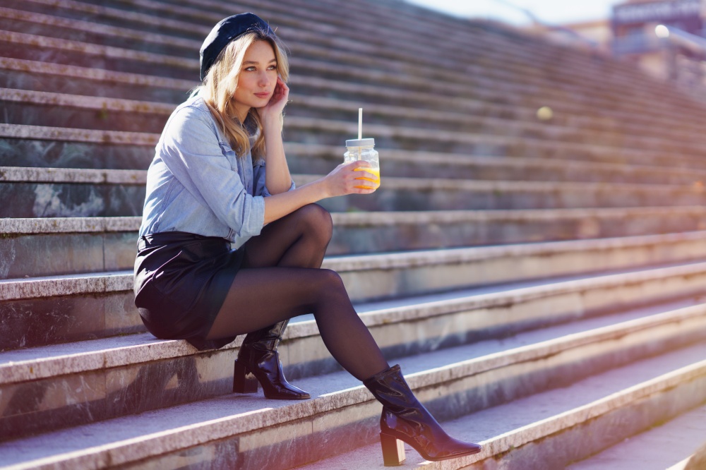 Blonde woman with beret, drinking a natural orange juice in a crystal glass, sitting on some urban steps.. Blonde woman with beret, drinking a natural orange juice in a crystal glass, sitting on some steps.