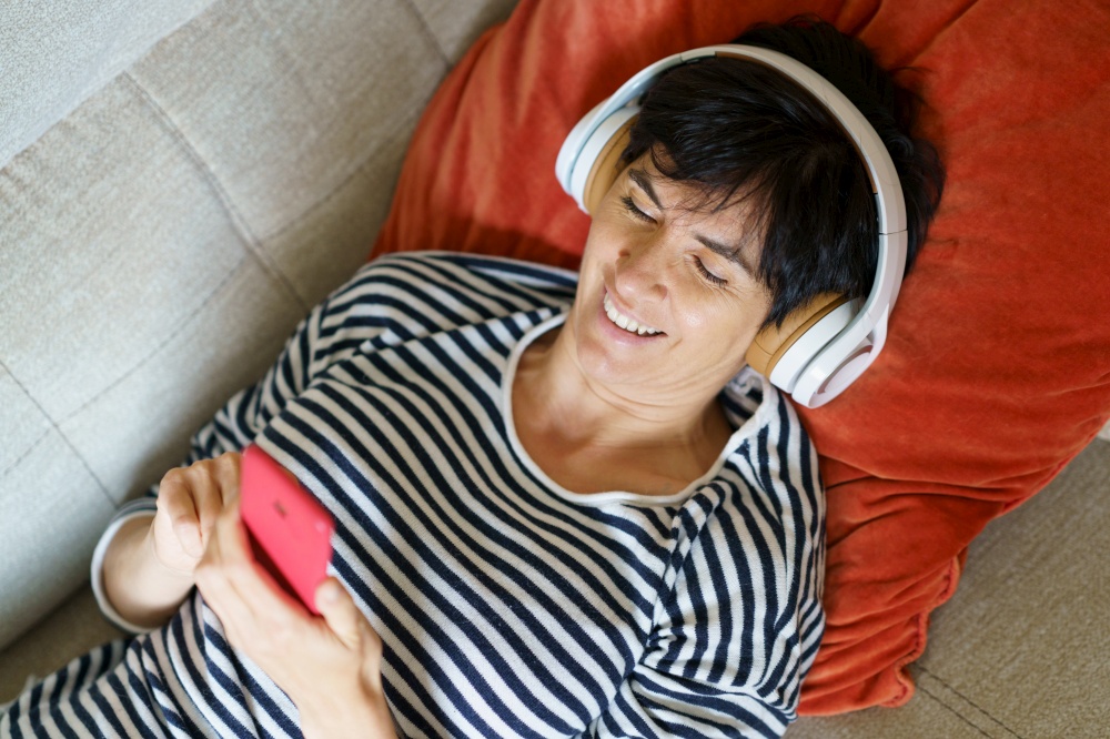 Woman smiling looking at her smartphone while listening to music with headphones lying on the sofa.. Woman smiling looking at her smartphone while listening to music with headphones.