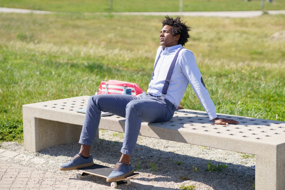 Black working man with afro hair taking a coffee break sitting on a park bench with his feet on his skateboard.. Black man with afro hair taking a coffee break sitting on a park bench.