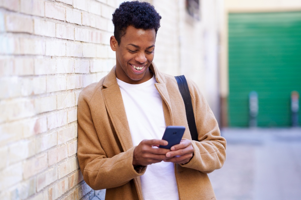 Young black man typing on his smartphone leaning against a brick wall in the street. Cuban man on urban background.. Young black man typing on his smartphone leaning against a brick wall in the street.
