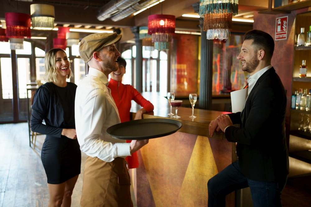 Male bartender with tray serving refreshing alcohol drinks on wooden counter for group of elegant people celebrating reunion in luxury bar. Barman serving cocktail on counter for clients
