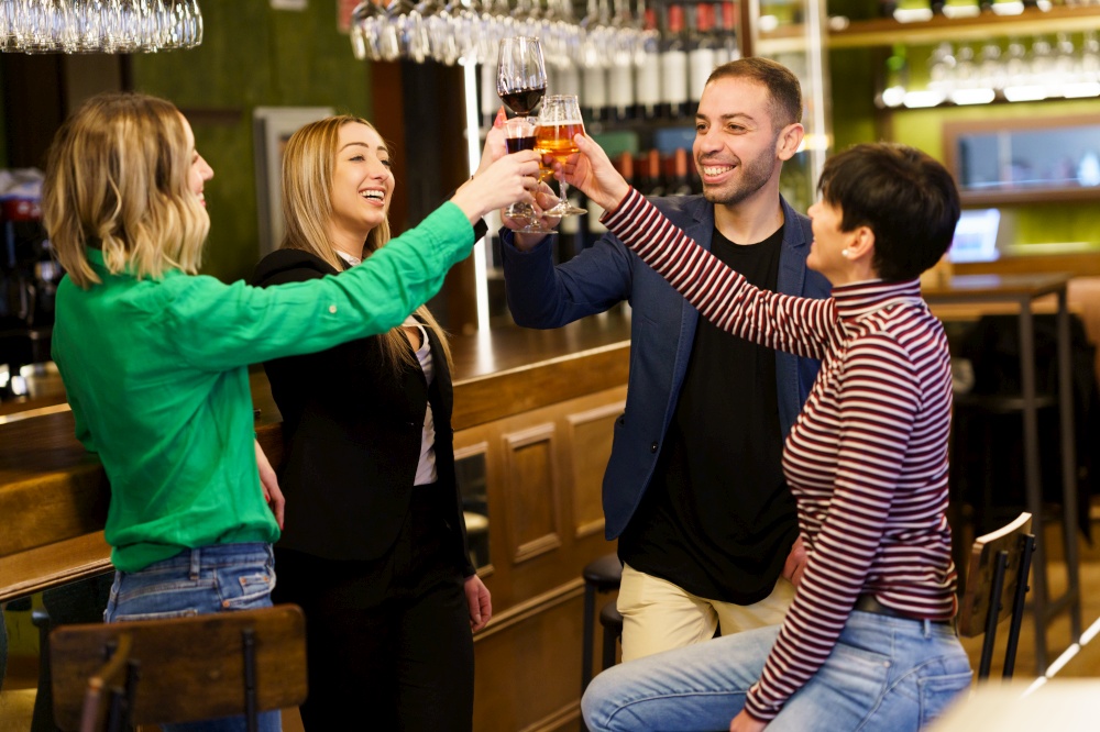 Merry multiethnic adult group of friends in casual clothes smiling and clinking glasses of alcohol drinks while resting near counter in weekend in bar. Happy young people proposing toast in a pub