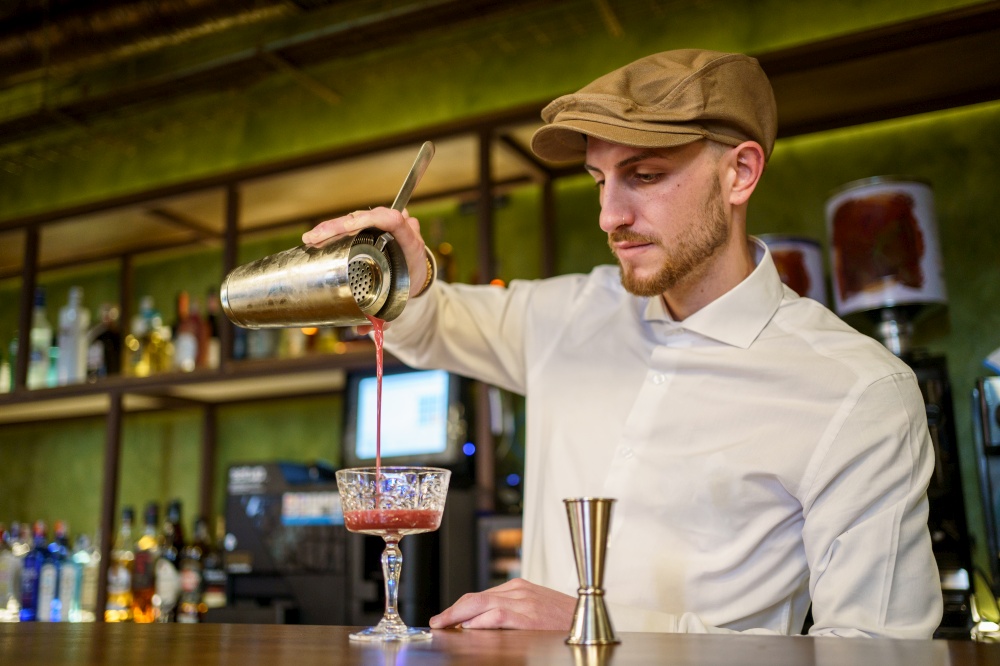 Low angle of bearded man in white shirt and cap filling glass goblet with fruit cocktail while working in bar in evening. Barman pouring cocktail into glass