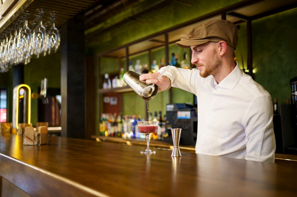 Bearded man in white shirt and cap filling glass with cocktail from shaker while standing behind counter and working in bar. Bearded bartender preparing cocktail in pub