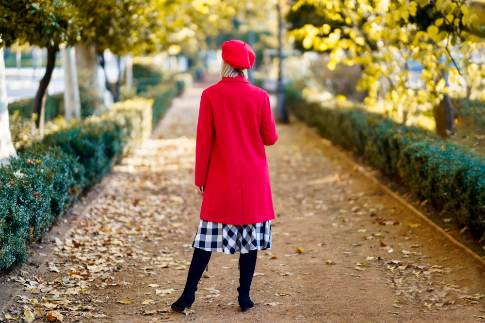 Full body back view of anonymous female in red outfit standing on pathway in park with trees on autumn day. Unrecognizable stylish woman in an urban park