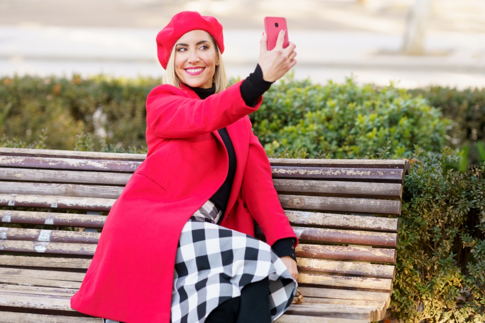 Delighted female in stylish red outfit taking self portrait on smartphone while sitting on wooden bench in park on autumn day. Cheerful woman taking selfie in park