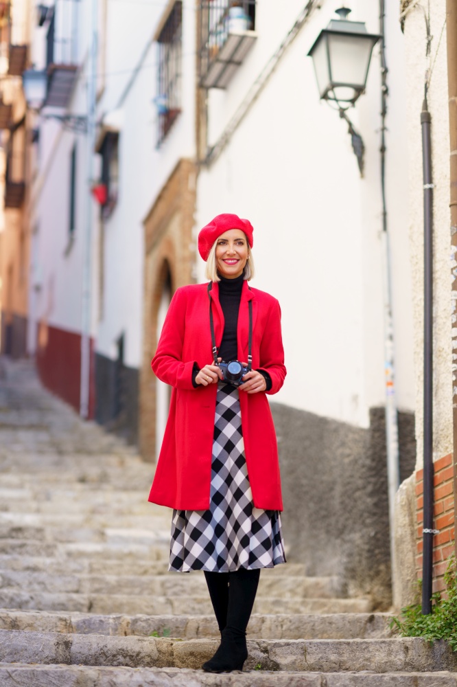 Full body of confident young female, tourist in fashionable outfit with photo camera, standing on old stairs in town and smiling while exploring city historic district in daytime. Fashionable lady standing on steps near aged building during vacation
