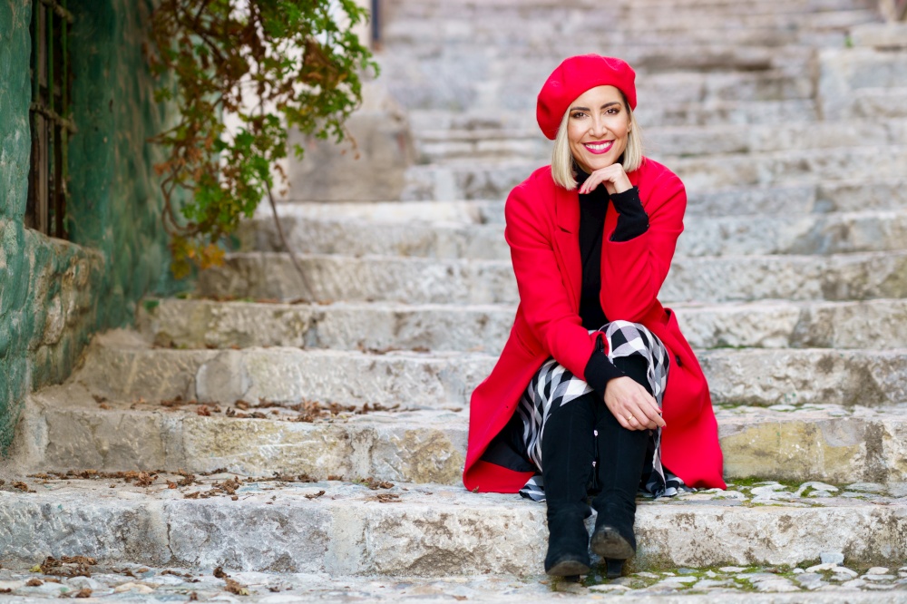 Full body of confident young lady, with blond hair and red lips in elegant coat and beret, sitting on shabby stone stairs with hand at chin and smiling in town. Feminine stylish woman resting on steps in town and smiling