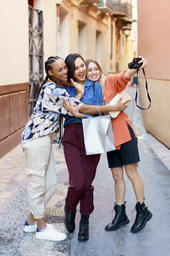 Full length of glad multiracial female friends taking selfie on camera while standing on street near building after shopping. Cheerful diverse ladies taking selfie portrait in town